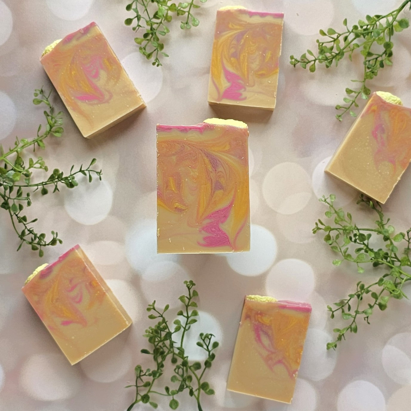 Six bars of Amillia Acres Pink Lemonade Goat Milk Soap are arranged on a light bokeh background, accented with small green leafy sprigs for decoration.