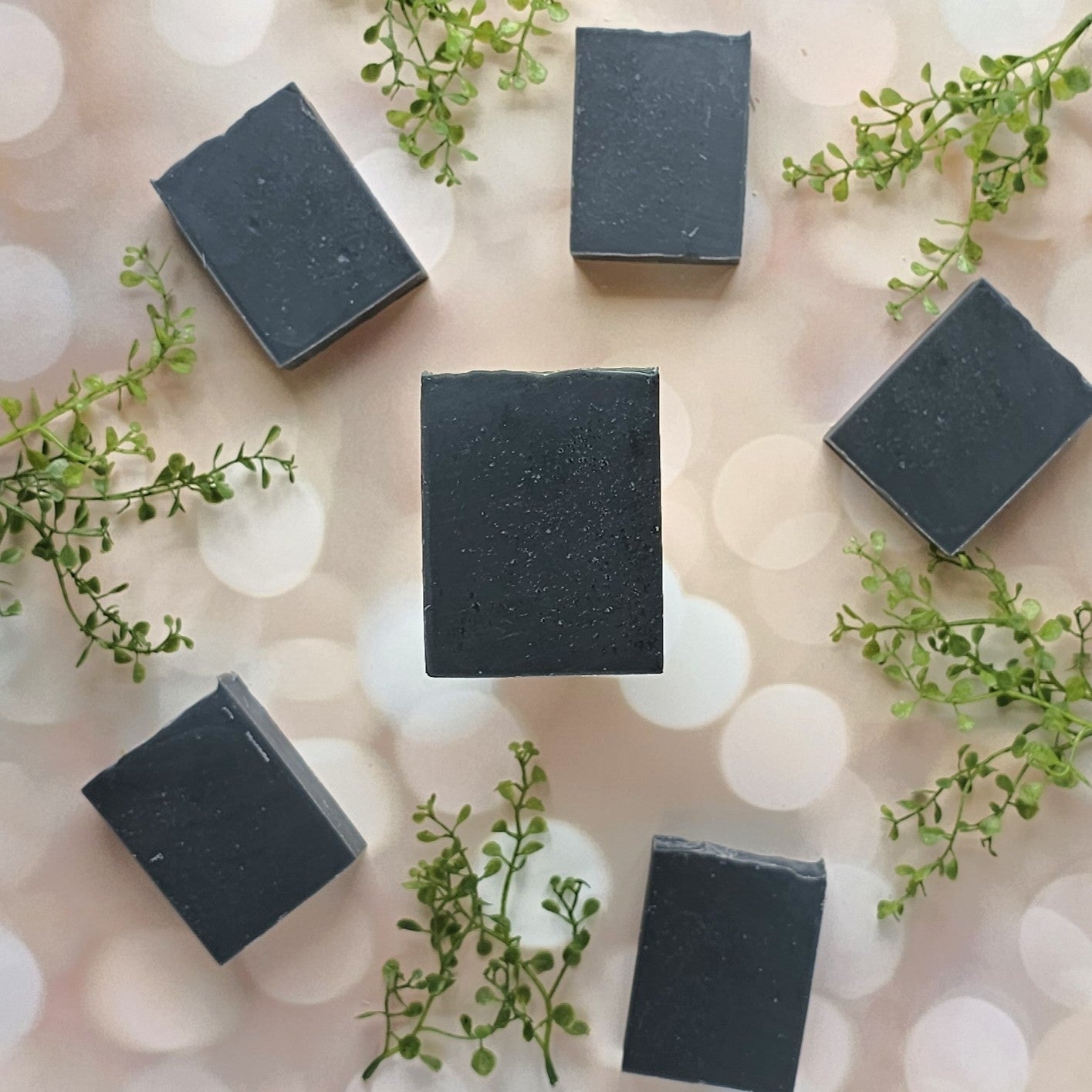 Six rectangular black Balance goat milk soap bars from Amillia Acres are arranged in a circle with green sprigs between them, set on a softly blurred light background featuring bokeh effects.