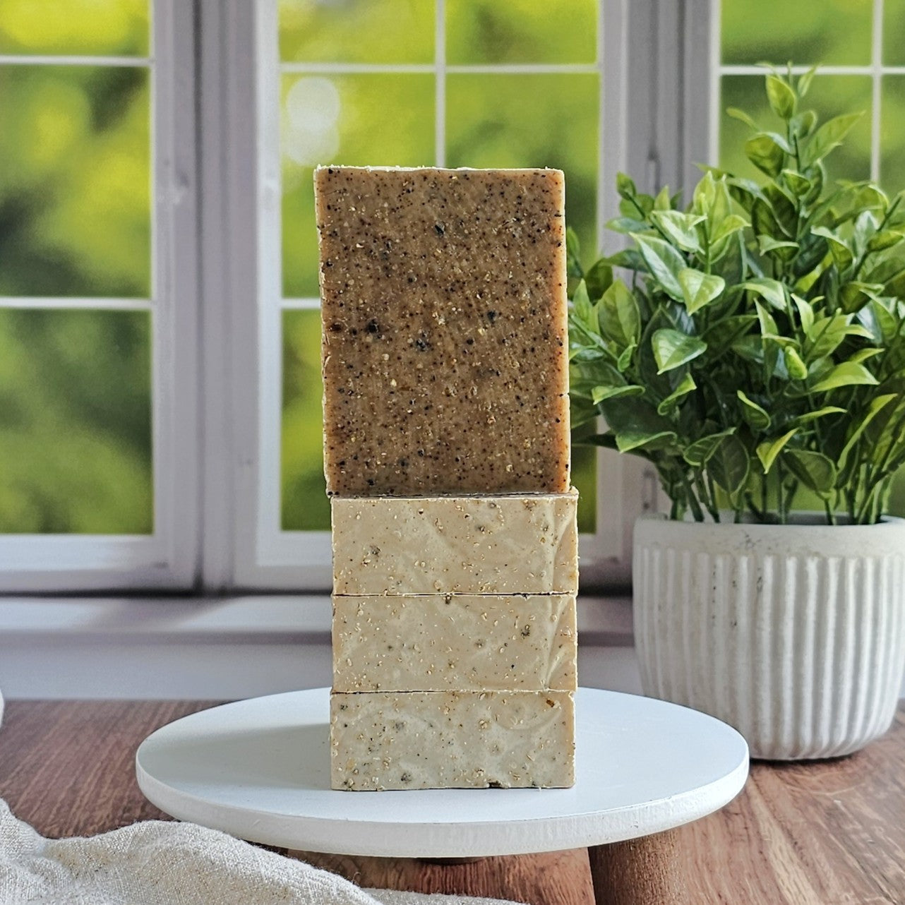 Three bars of Gardener Goat Milk Soap by Amillia Acres ~ Goat Milk Soaps and Lotions are stacked on a white pedestal near a window with greenery outside, alongside a potted plant for a fresh, natural touch.
