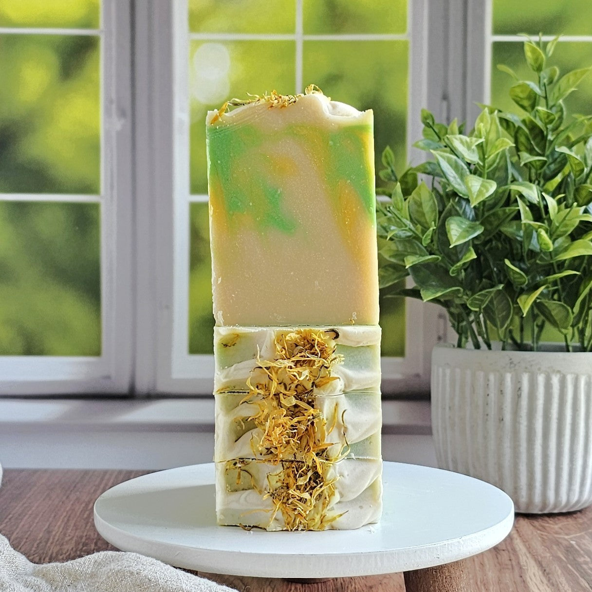 Two handmade soaps from Amillia Acres ~ Goat Milk Soaps and Lotions, featuring the Jasmine & Honeysuckle goat milk soap with swirled green, yellow, and cream colors, are stacked on a white stand and decorated with dried yellow flowers by a window.