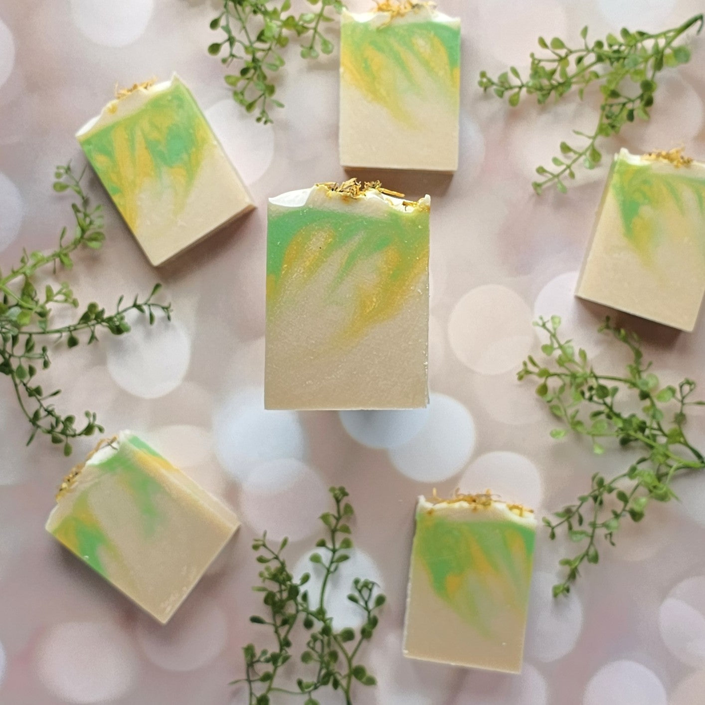 Six Jasmine & Honeysuckle Goat Milk Soaps from Amillia Acres are arranged in a circle on a soft bokeh background, each featuring green and yellow swirls and sprigs of greenery between them.