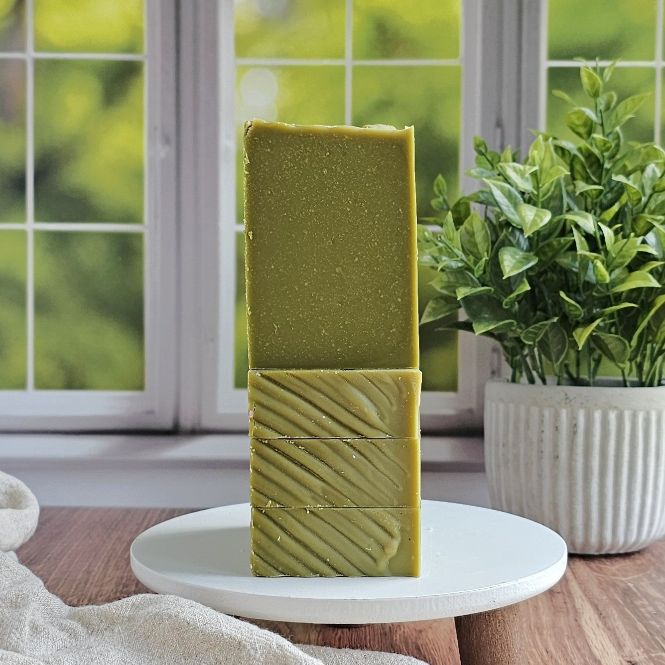 Two green Minty (Essential Oil) Goat Milk Soap bars from Amillia Acres are stacked on a white stand by a window with lush greenery. A plant in a white pot and a beige towel add to the clean, natural feel of the scene.
