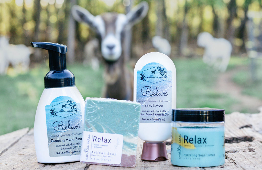 Various Relax fragranced products - foaming hand soap, bar soap, goat milk lotion, and sugar scrub on a wood table with goats blurred in the background