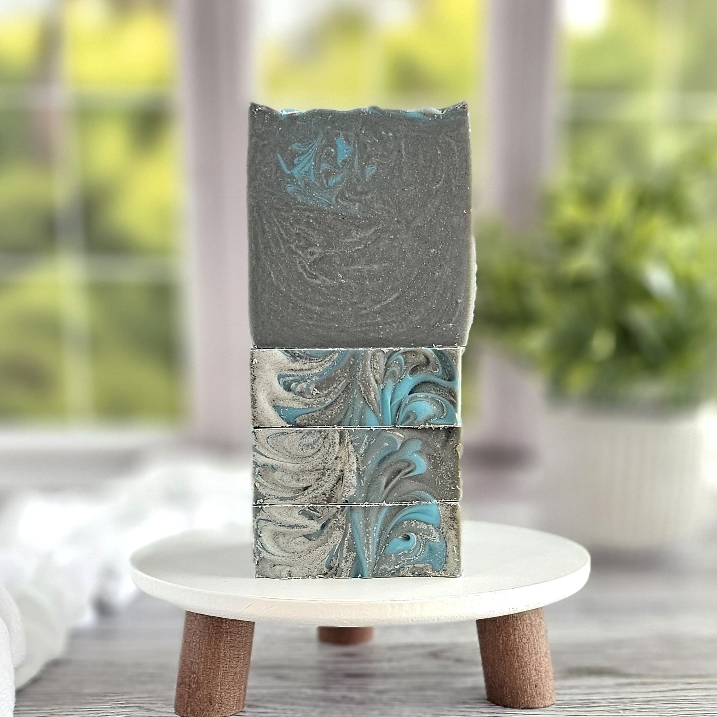Three Bay Rum Goat Milk Soaps by Amillia Acres, featuring gray and blue swirls, are elegantly stacked on a small white stand. Green foliage through a window adds nature's serene touch to this artisanal display.