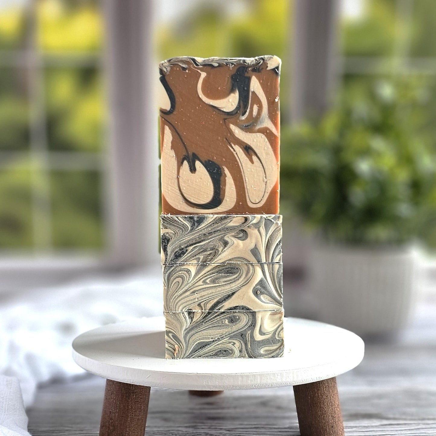Three "Man Cave" goat milk soaps by Amillia Acres, featuring intricate marbled patterns in brown, cream, and black, are stacked on a small stool. Infused with natural oils for a luxurious feel, the scene captures greenery through a window in the blurred background.