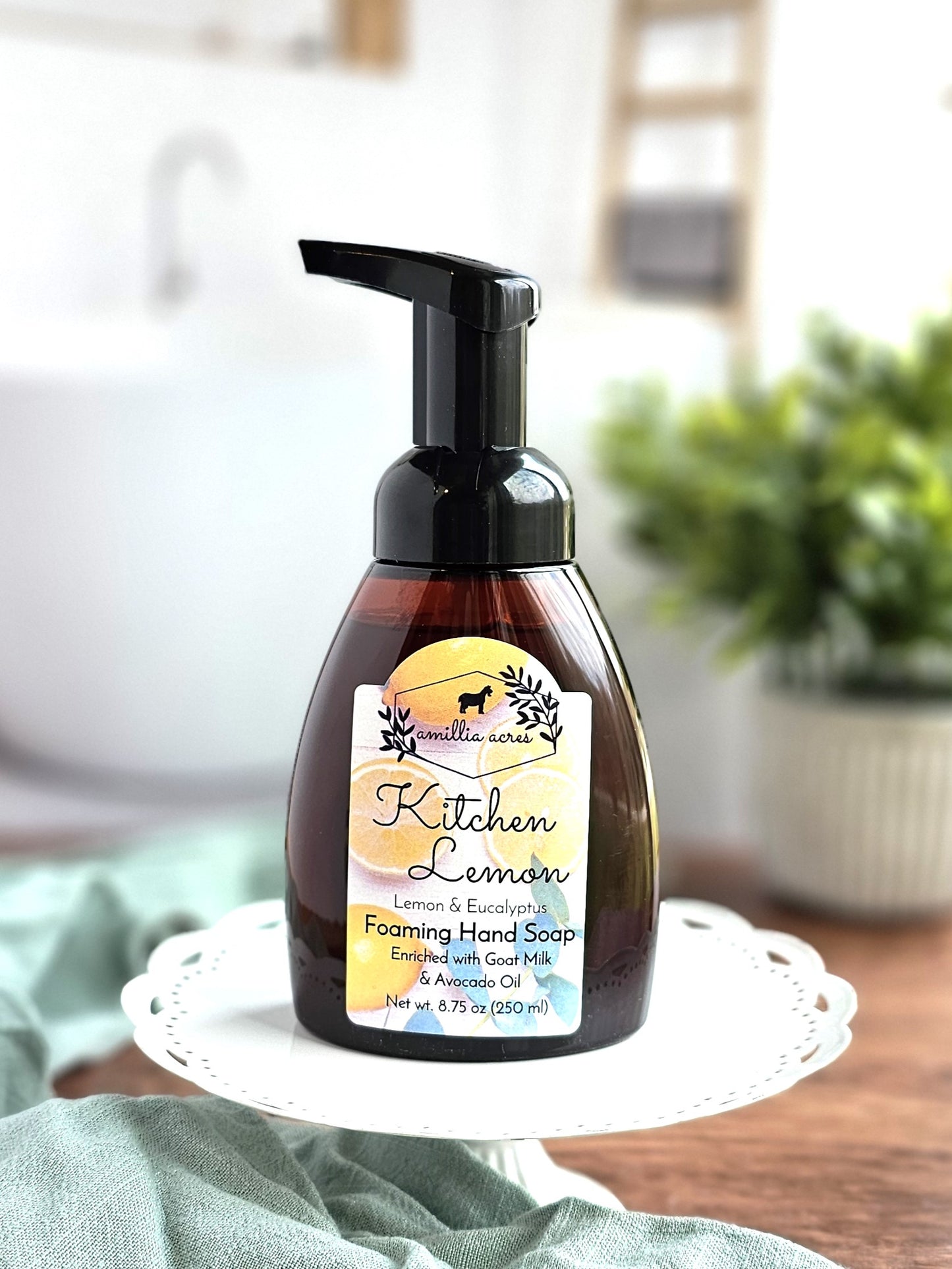 A brown bottle of Amillia Acres Foaming Hand Soap labeled "Kitchen Lemon" with moisturizing goat milk and avocado oil sits on a white decorative stand, featuring blurred plant and kitchen items in the background.