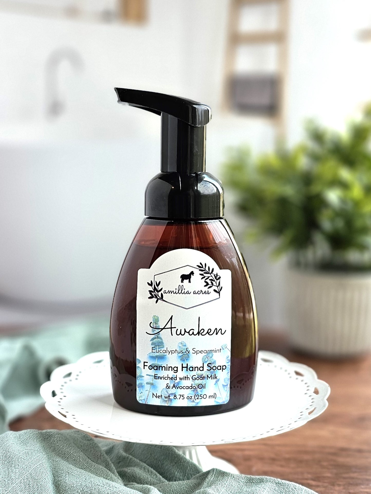 A bottle of Amillia Acres' Awaken foaming hand soap, scented with eucalyptus and spearmint, elegantly adorns a decorative stand. The label promotes its moisturizing goat milk and avocado oil blend. A blurred bathroom background with a plant enhances the serene setting.