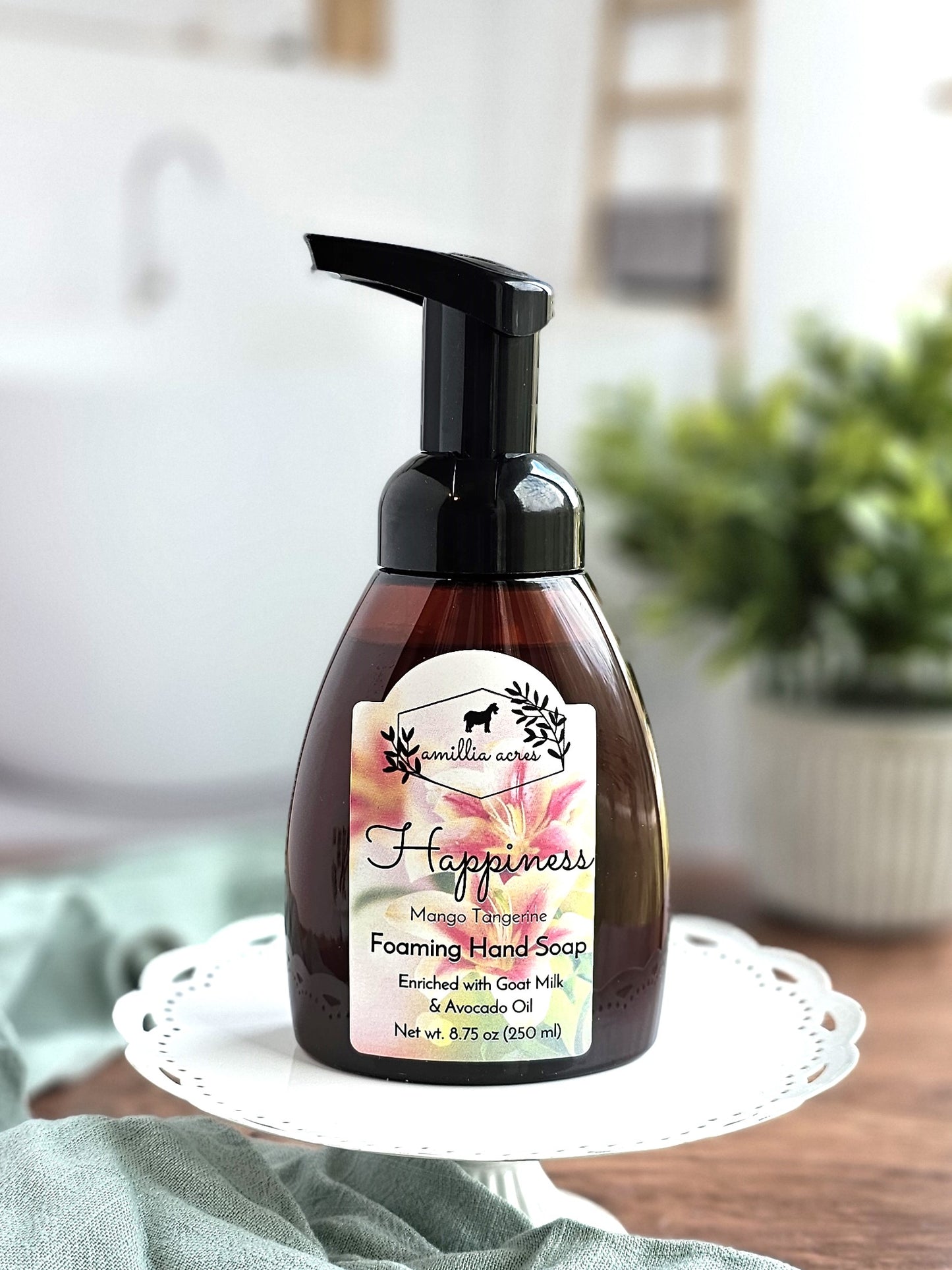 A bottle of Amillia Acres "Happiness" Foaming Hand Soap in Mango Tangerine rests on a white stand. The label highlights its moisturizing blend with goat milk and avocado oil. A plant and blurred indoor elements create a serene backdrop.