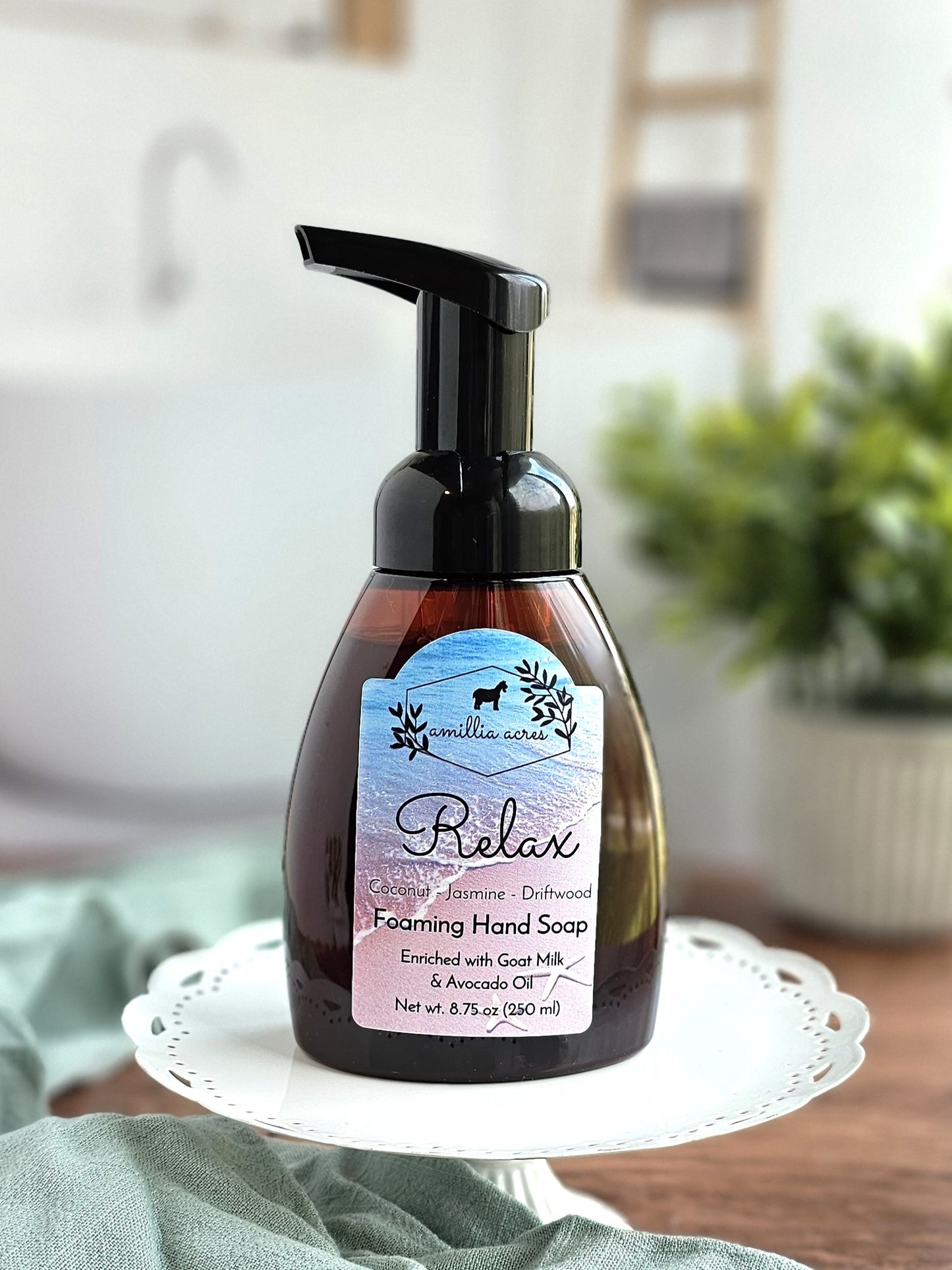 A pump bottle of Amillia Acres' "Relax" Foaming Hand Soap, featuring jasmine and driftwood on the label, enriched with goat milk and avocado oil. It sits elegantly on a decorative white plate with a softly blurred plant in the background.