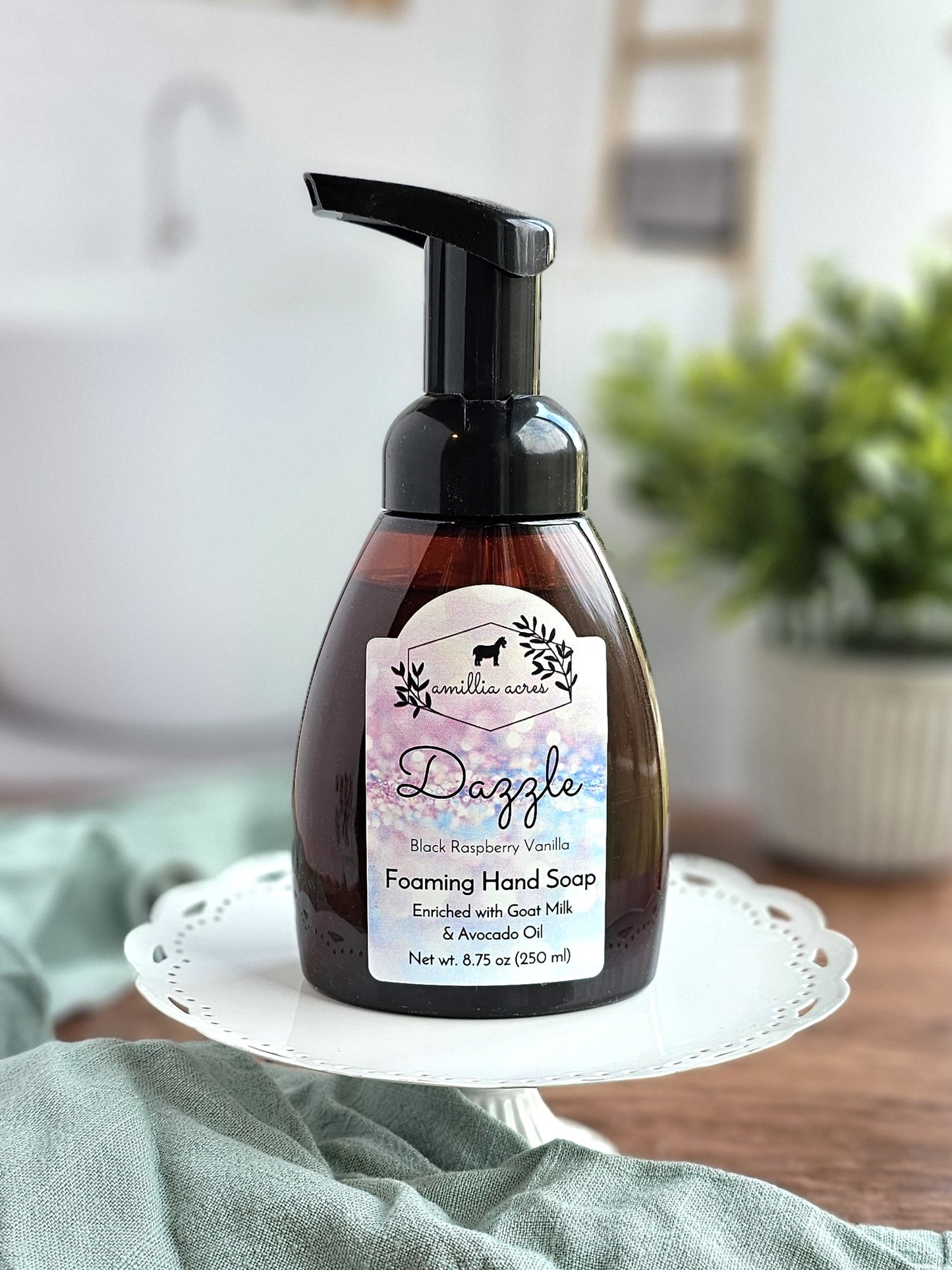 A Dazzle bottle of Amillia Acres' Black Raspberry Vanilla Foaming Hand Soap, celebrated for its moisturizing goat milk and avocado oil blend, sits elegantly on a white cake stand. A blurred plant enhances the charm alongside a rustic wooden piece in the background.