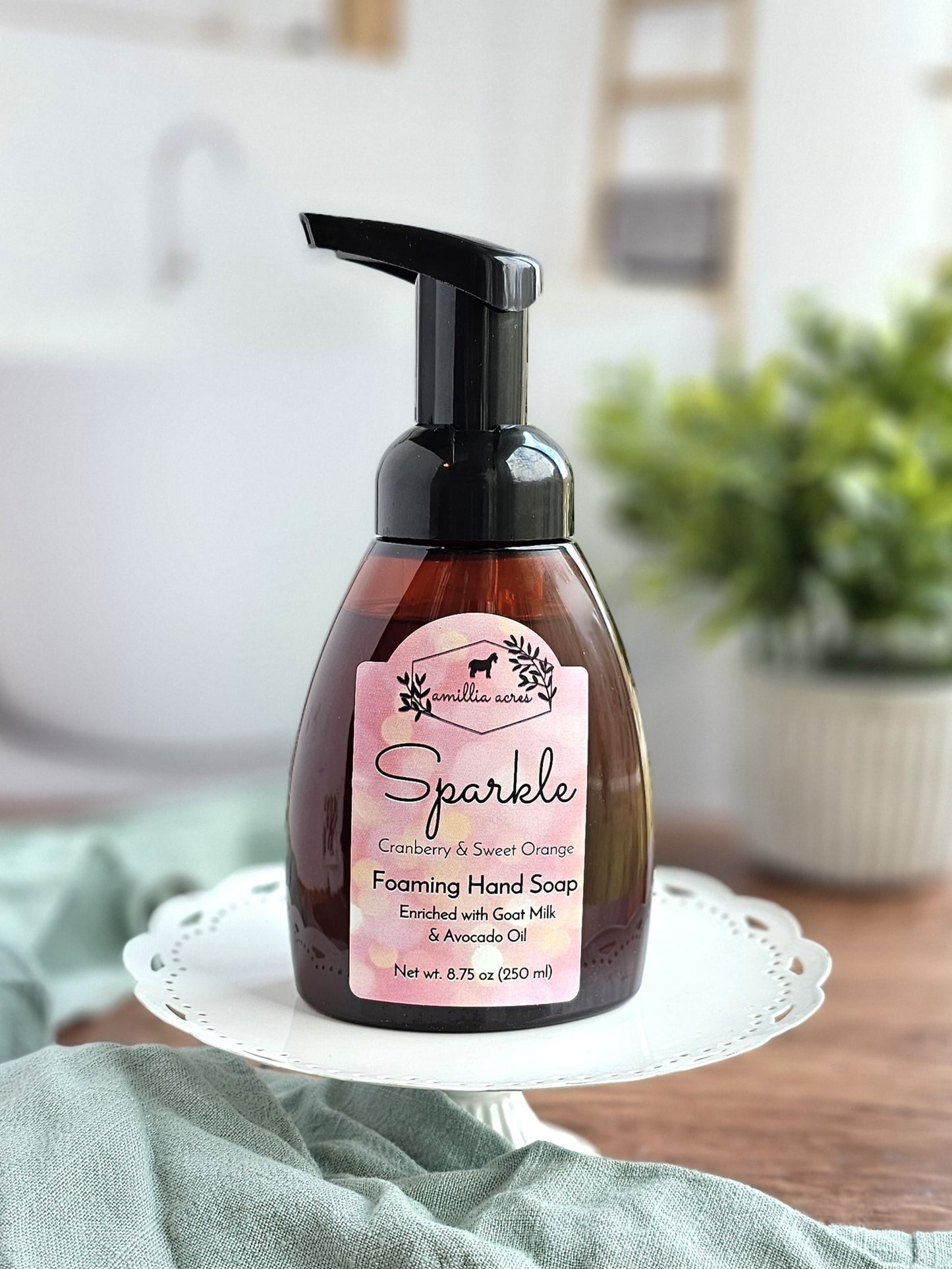 A pump bottle of "Sparkle" foaming hand soap by Amillia Acres, featuring cranberry and sweet orange scents, enriched with goat milk and avocado oil, rests on a white plate in a kitchen with a blurred green plant in the background.