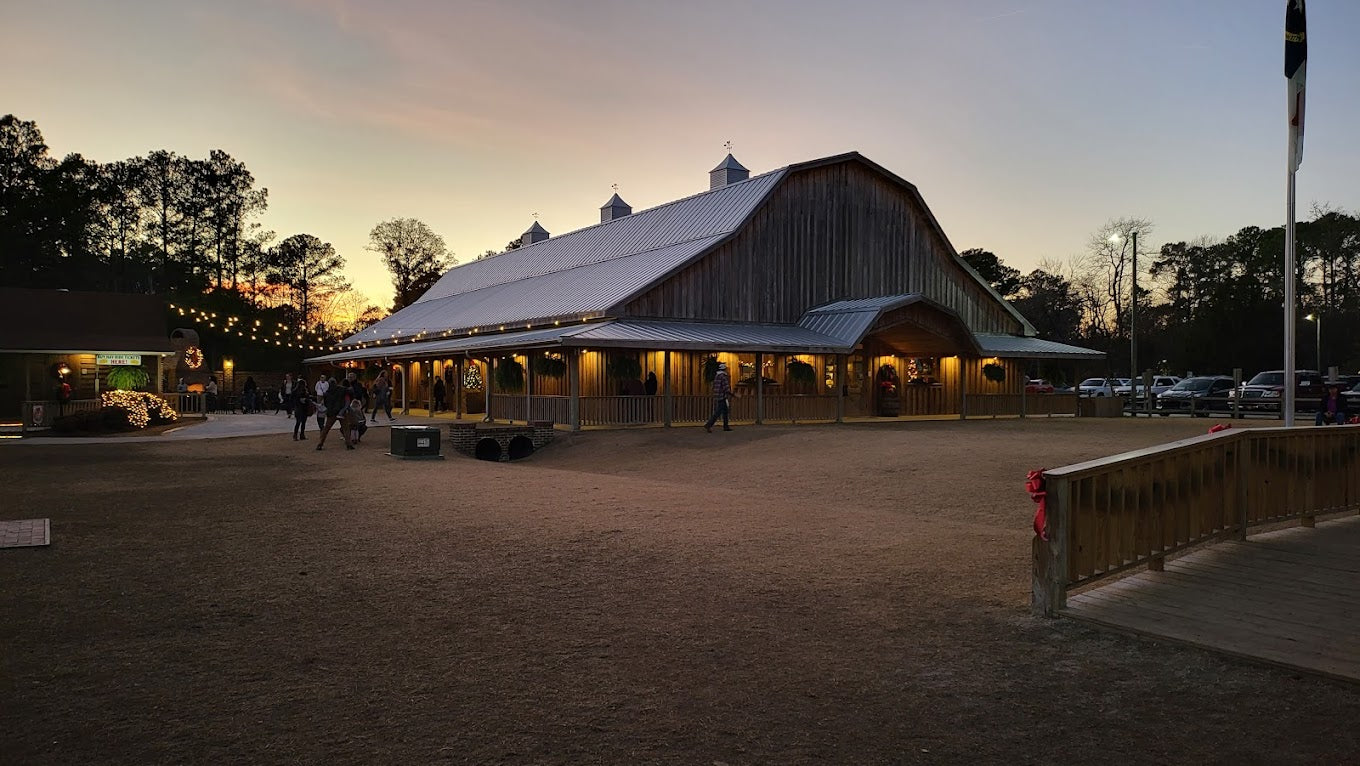A large wooden barn-like structure is lit up in the evening. People can be seen walking nearby, and surrounding trees are silhouetted against the sunset. The ambiance is calm and festive.