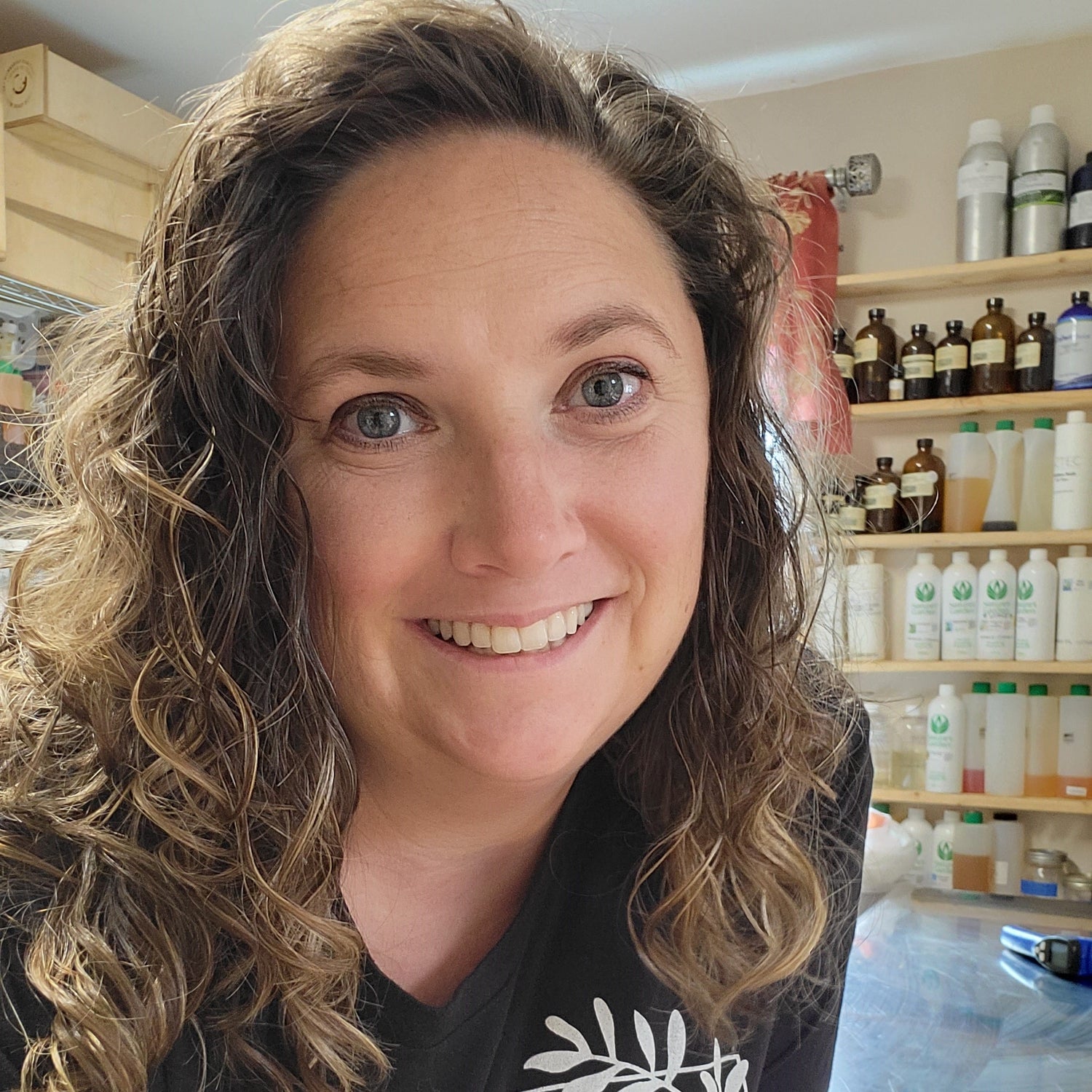 A person with curly hair smiles at the camera in a room filled with shelves stocked with bottles and containers.