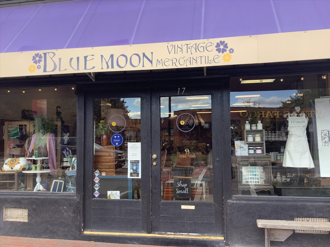 Street view of "Blue Moon Vintage Mercantile" store with a purple awning. Display window shows vintage items like clothing and decor. A "Shop Small" sign is on the door, and interior shelves are visible through the glass.