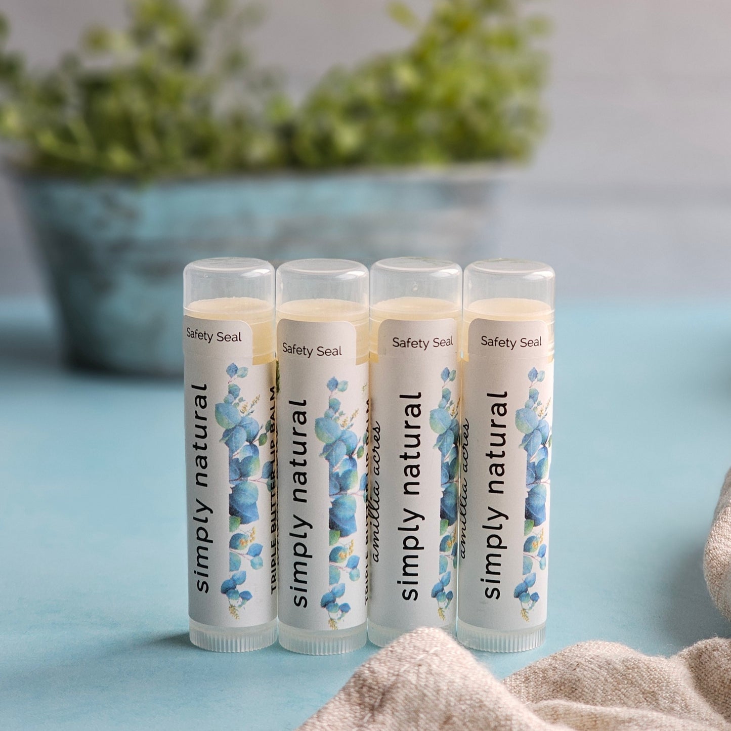Four tubes of Amillia Acres' "Natural | Triple Butter Lip Balm," adorned with a floral design, are upright on blue. Enriched with antioxidants, these balms rejuvenate lips. A blurred green plant and beige cloth appear in the background.