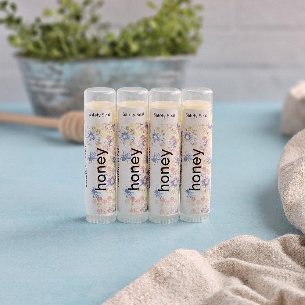 Four tubes of Honey | Triple Butter Lip Balm by Amillia Acres ~ Goat Milk Soaps and Lotions, featuring floral designs, are arranged on a light blue surface. A honey dipper, green leaves, and a beige cloth enhance the luxurious moisturizing effect of these nourishing balms.