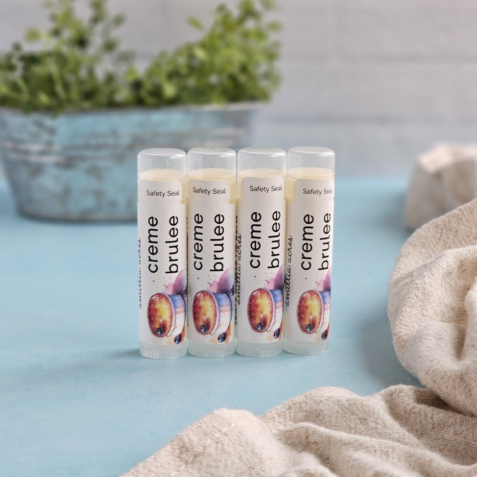 Four Creme Brulee | Triple Butter Lip Balm tubes by Amillia Acres ~ Goat Milk Soaps and Lotions stand on a light blue surface, crafted with nourishing Mango Butter. A rustic pot with greenery is blurred in the background, while beige fabric peeks from the right.