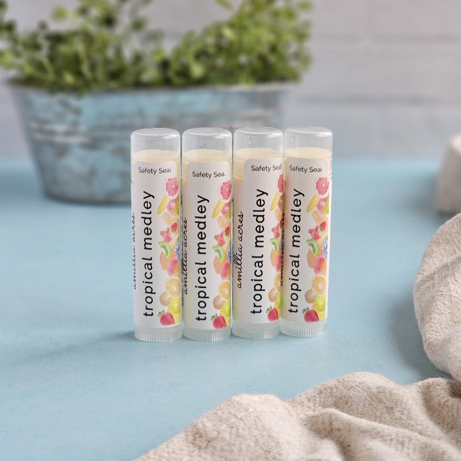 Four Tropical Medley lip balms from Amillia Acres ~ Goat Milk Soaps and Lotions stand upright on a blue surface, featuring vibrant fruit illustrations. Their antioxidant-rich triple butter formula provides colorful nourishment. A blurred potted plant and beige cloth add to the serene scene.