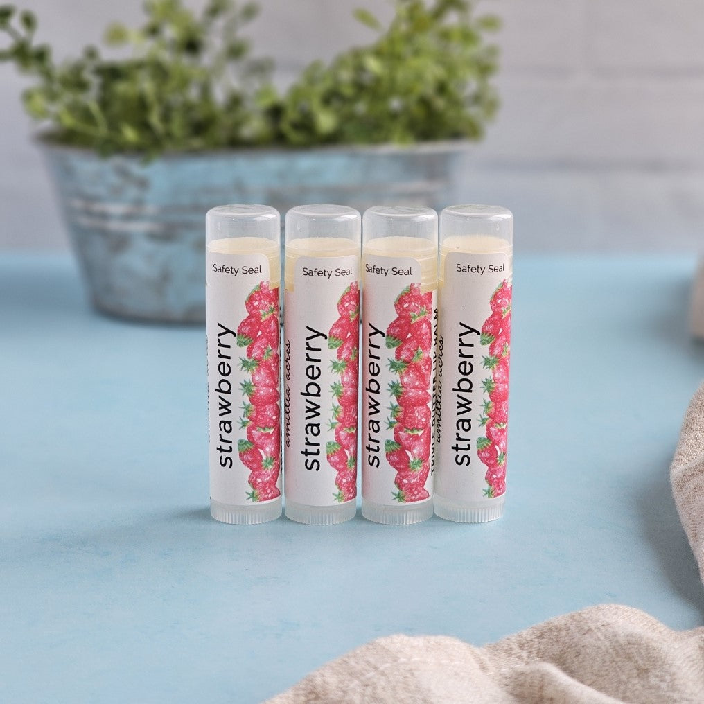 Four tubes of Amillia Acres ~ Goat Milk Soaps and Lotions' Strawberry Triple Butter Lip Balm stand upright on a light blue surface. Each tube features "strawberry" labels with strawberry images, using a nourishing recipe. A blurred potted plant is in the background.