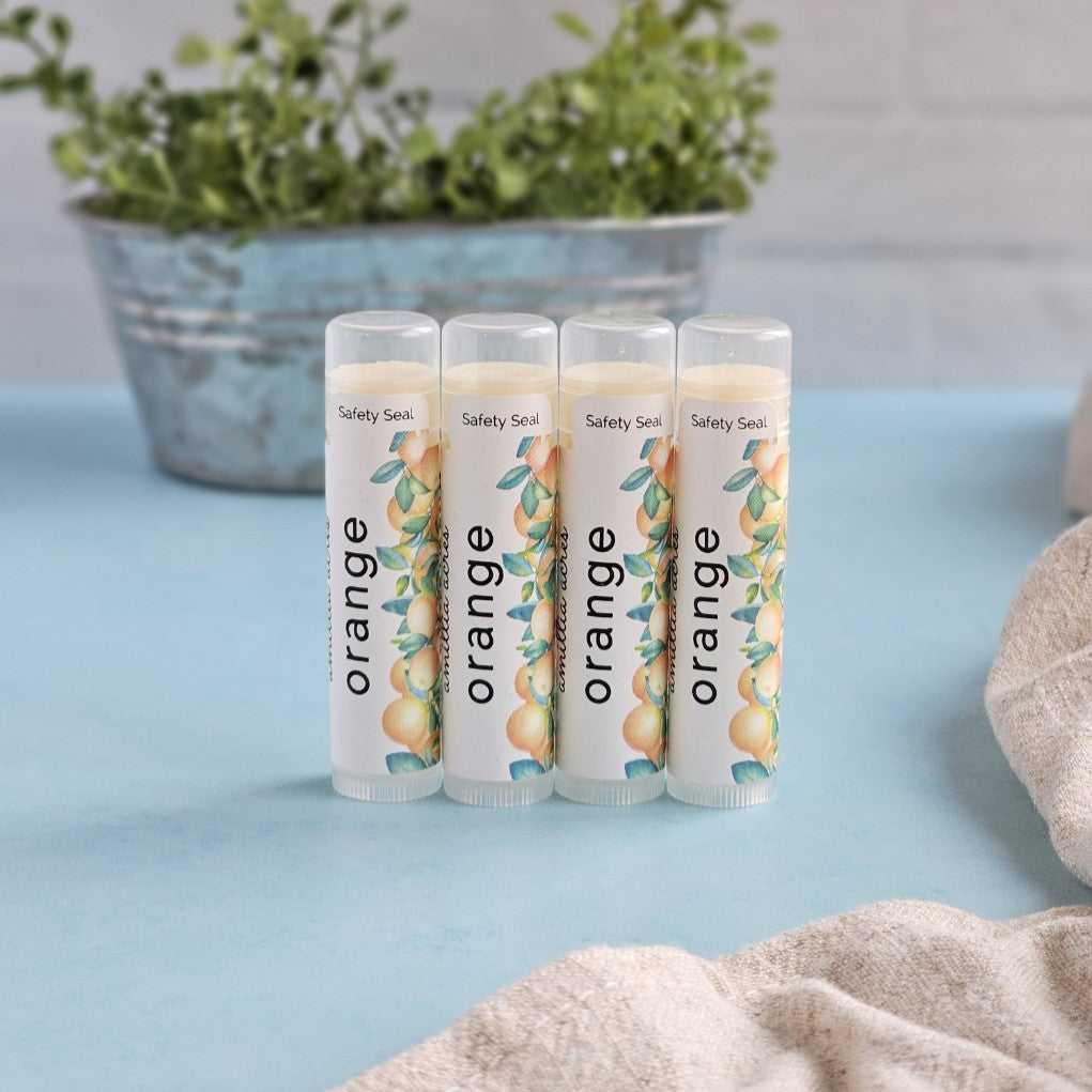 Four tubes of Amillia Acres' Orange Triple Butter Lip Balm are lined up on a blue surface, each featuring "orange," illustrations of oranges, leaves, and antioxidants. A blurred plant is in the background with a beige cloth partially visible in the foreground.