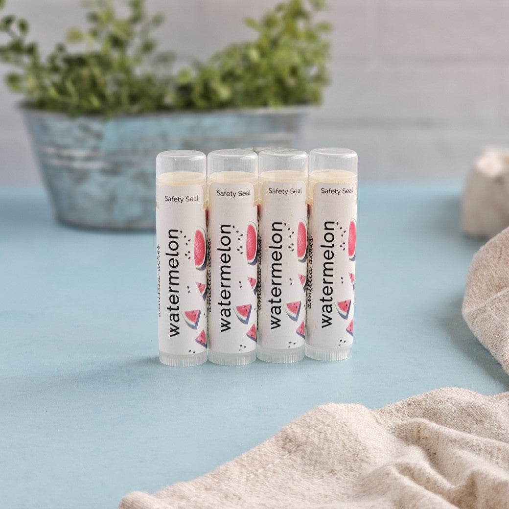 Four tubes of Amillia Acres Watermelon Triple Butter Lip Balm enriched with antioxidants stand on a light blue surface. Each tube has watermelon illustrations and a safety seal. A blurred metal planter with greenery is in the background, with a beige cloth partially visible.