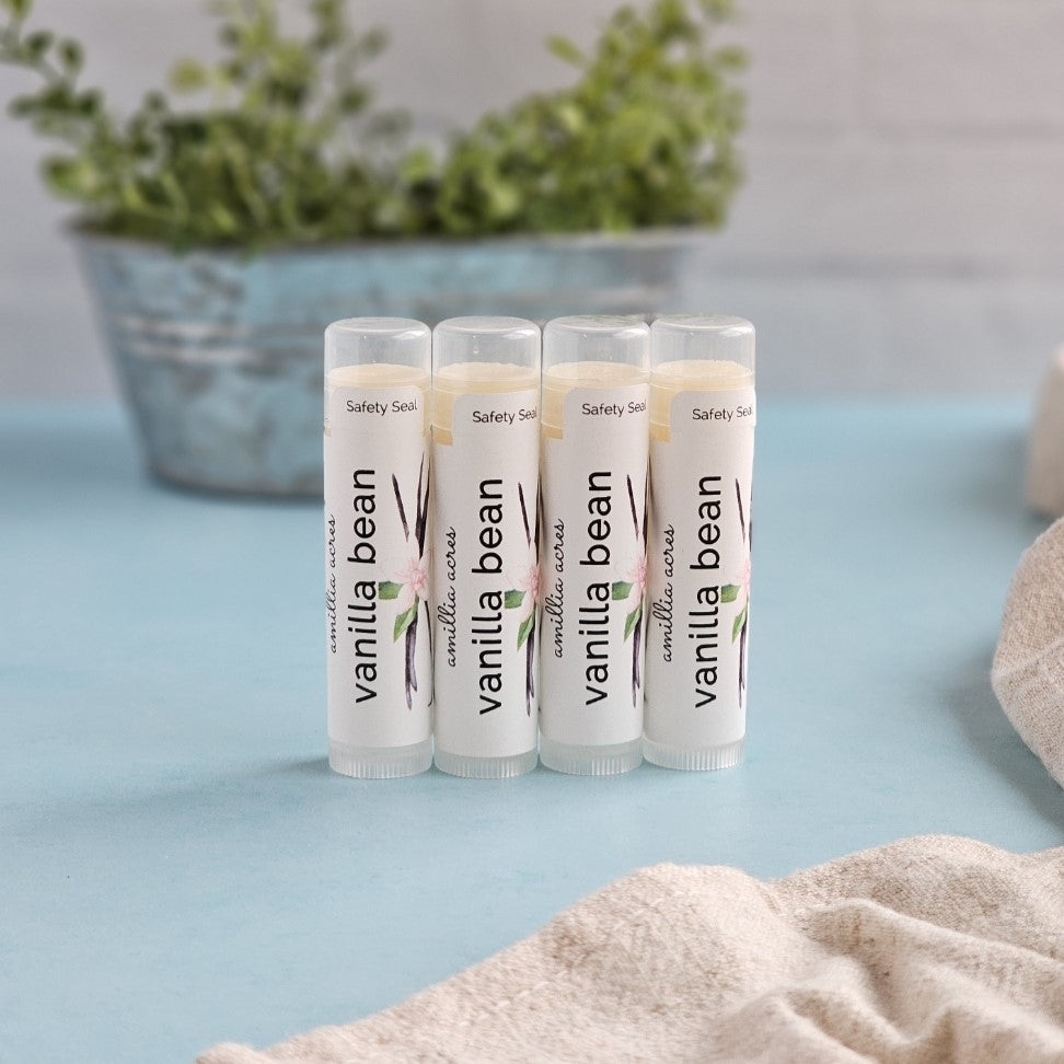 Four tubes of Vanilla Bean Triple Butter Lip Balm by Amillia Acres ~ Goat Milk Soaps and Lotions are on a light blue surface. A metallic pot with green foliage and beige fabric in the background creates a cozy, natural setting enriched with antioxidants to enhance their nourishing recipe.