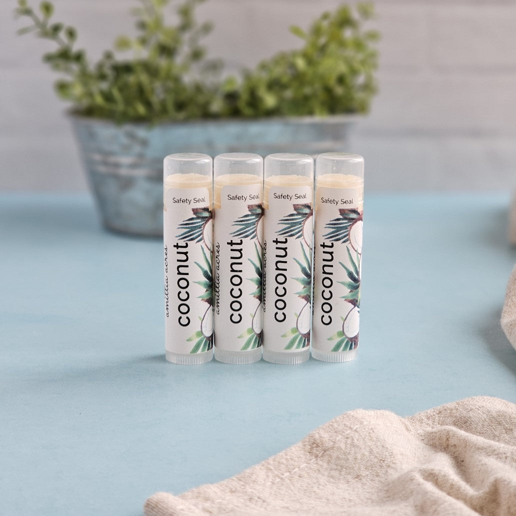 Four tubes of Amillia Acres ~ Goat Milk Soaps and Lotions' "Coconut | Triple Butter Lip Balm" sit on a light blue surface, adorned with coconut illustrations. A plant in a metal container stands behind them, with part of a cloth peeking from the side.
