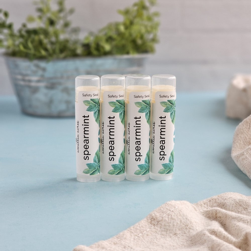 Four tubes of Amillia Acres Spearmint Triple Butter Lip Balm are arranged on a light blue surface with a blurred plant in a metallic pot in the background. A beige cloth is partially visible on the right, enhancing the serene setting.