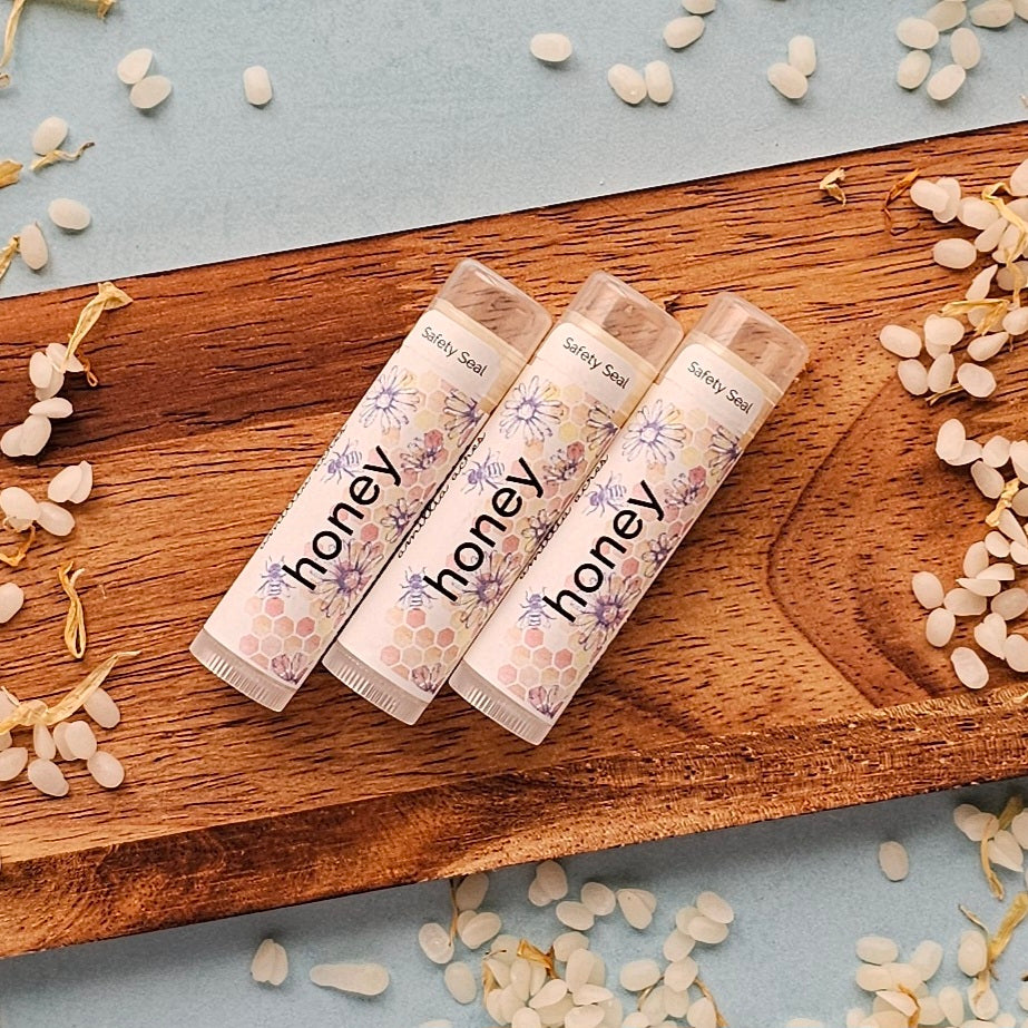 Three Honey | Triple Butter Lip Balms from Amillia Acres ~ Goat Milk Soaps and Lotions, featuring floral designs, rest on a wooden tray. Enriched to moisturize lips, they are surrounded by small white beads and dried flowers on a light blue surface.