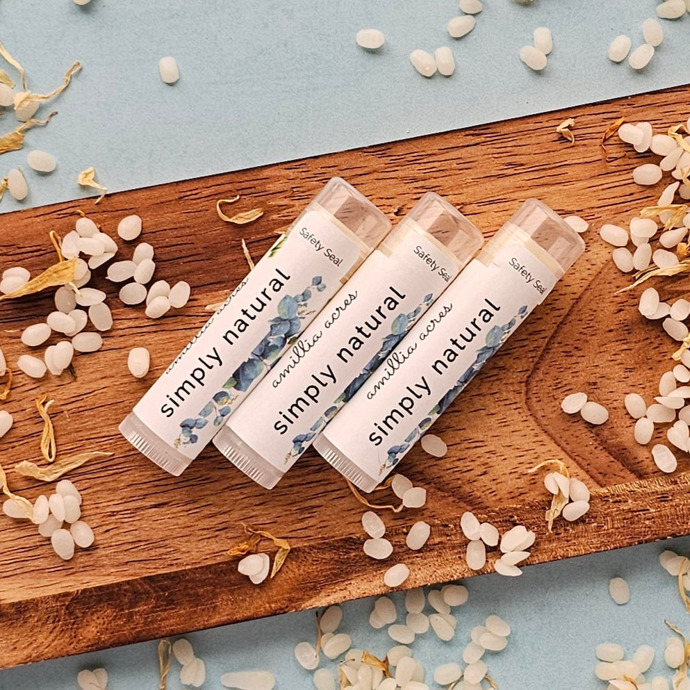 Three tubes of Amillia Acres' "Natural" Triple Butter Lip Balm are displayed on a wooden board, crafted with a nourishing recipe rich in antioxidants. Small white beads and petals are scattered around on a light blue surface, adding an elegant touch.