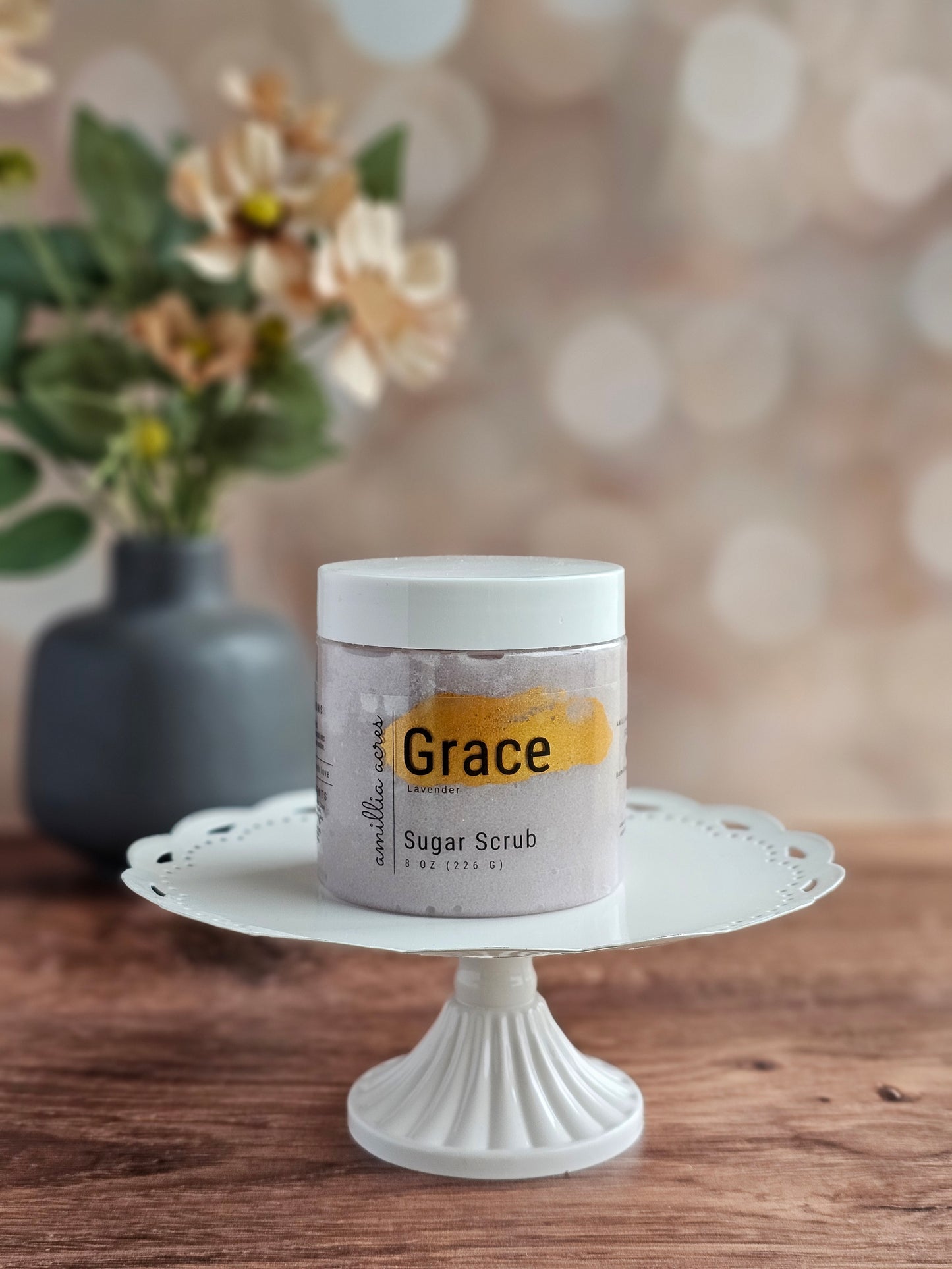 A jar of Amillia Acres ~ Goat Milk Soaps and Lotions Grace Sugar Scrub rests on a white decorative cake stand atop wood, highlighting this luxurious exfoliating body scrub with a blurred floral vase in the background.