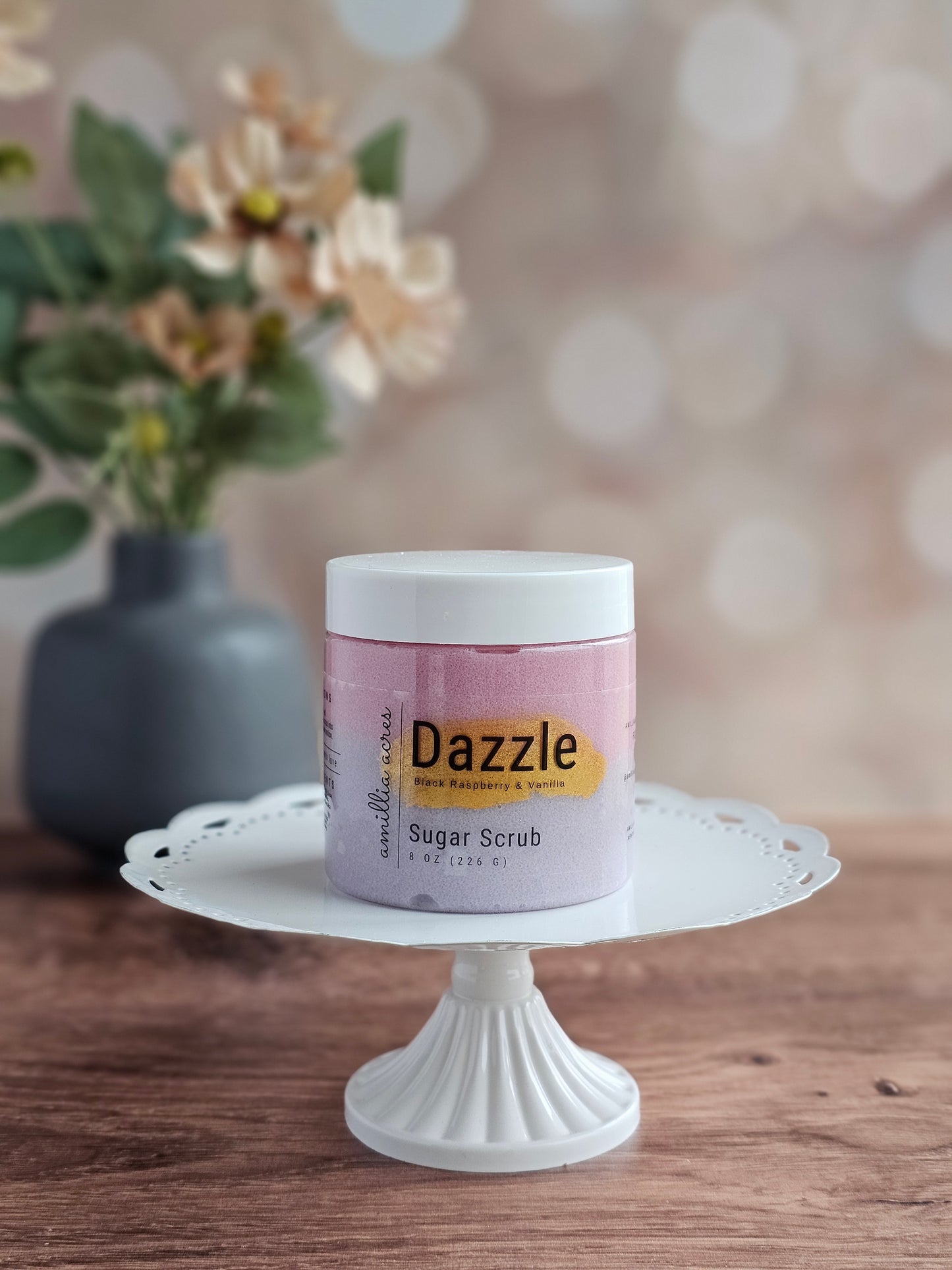 A jar of Dazzle Sugar Scrub by Amillia Acres ~ Goat Milk Soaps and Lotions in Black Raspberry Vanilla rests on a white cake stand on a wooden surface, with blurred flowers visible in the background.