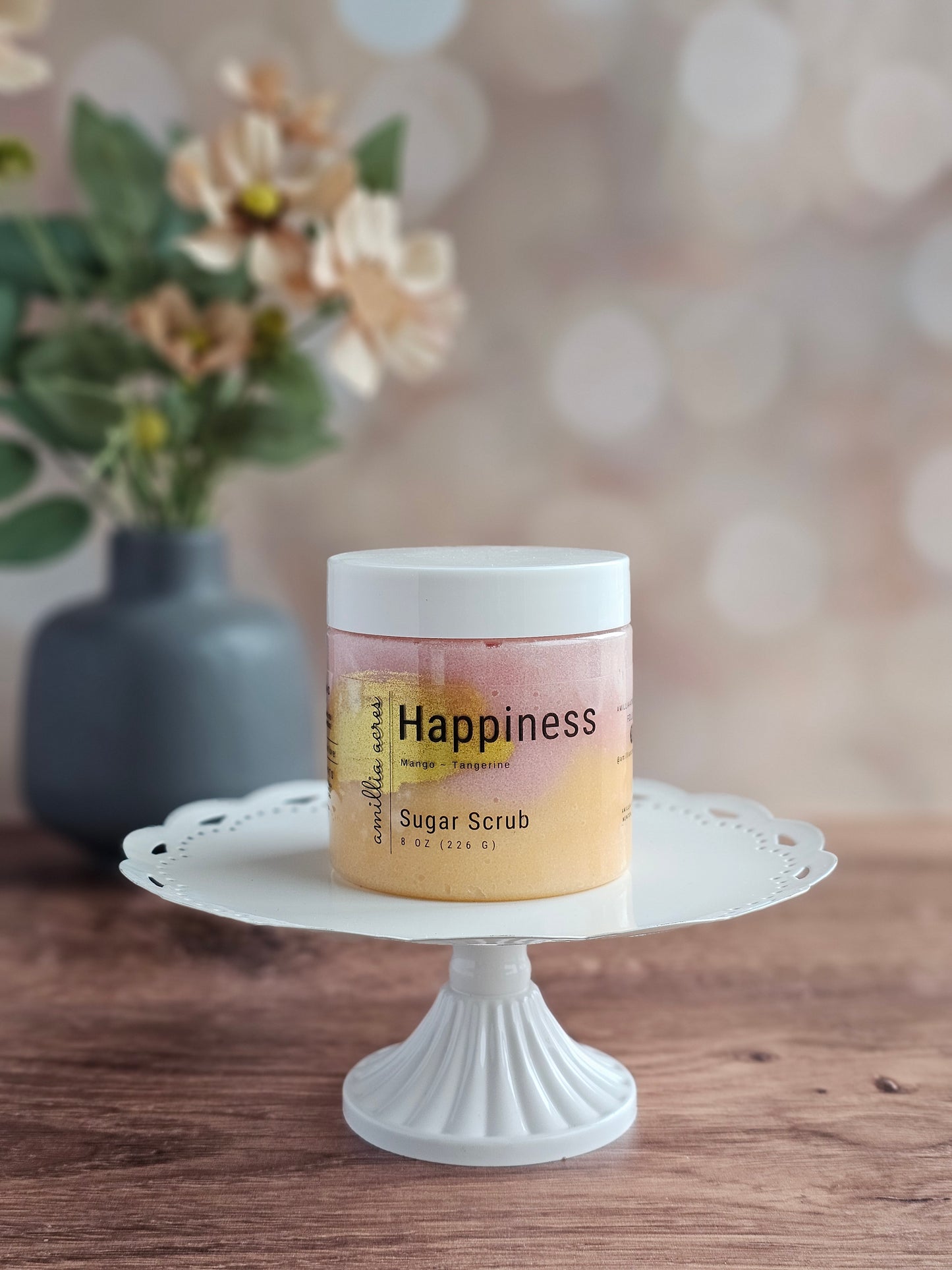 A jar of Happiness Sugar Scrub by Amillia Acres ~ Goat Milk Soaps and Lotions sits on a white cake stand, offering glowing skin with its exfoliating blend; a blurred vase of flowers rests in the background on a wooden surface.