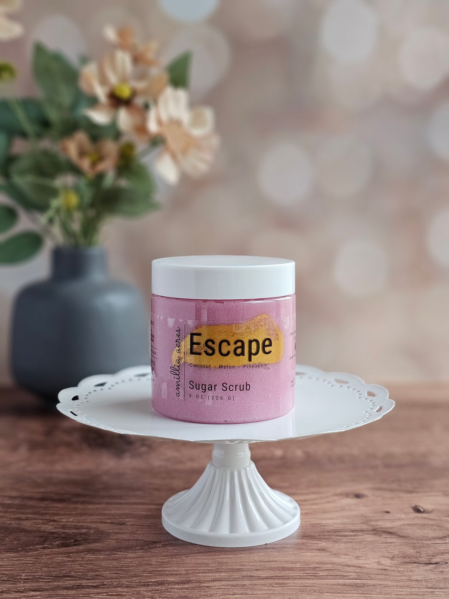 A pink jar of Amillia Acres ~ Goat Milk Soaps and Lotions "Escape | Sugar Scrub" exfoliating body scrub for glowing skin sits on a white cake stand, with a blurred gray vase of flowers in the background on a wooden surface.