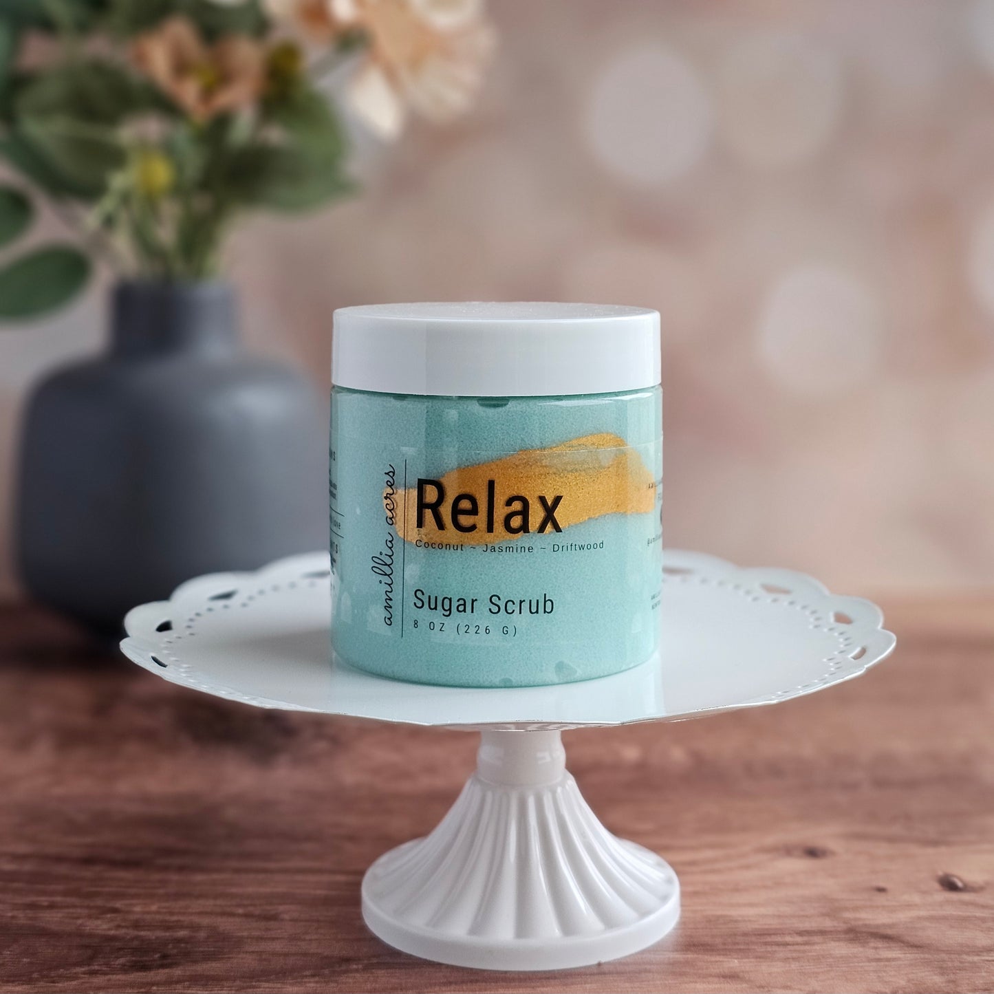 A jar of Amillia Acres ~ Goat Milk Soaps and Lotions "Relax | Sugar Scrub" rests on a white pedestal, with a soft, floral background—showcasing this exfoliating body scrub for glowing skin.