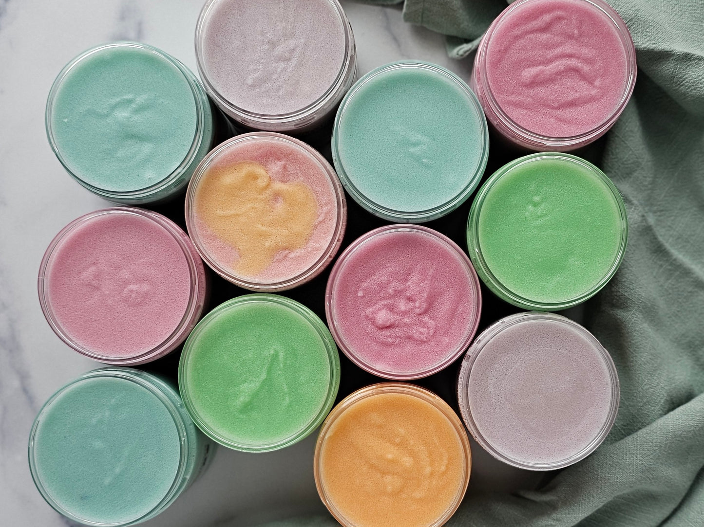 A top view of twelve jars of Amillia Acres ~ Goat Milk Soaps and Lotions' Happiness Sugar Scrub in bright pink, green, blue, and yellow, arranged neatly on a marble surface next to soft green fabric.