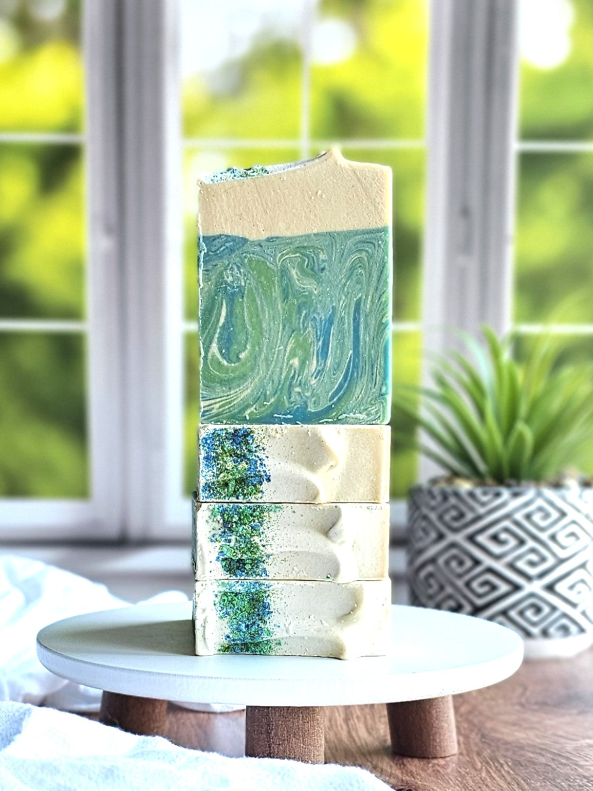 Three "Awaken" goat milk soaps by Amillia Acres, swirled in blue and white with eucalyptus and spearmint scents, rest on a round wooden stand. A plant in a patterned pot complements the tranquil scene with a blurred green window view.