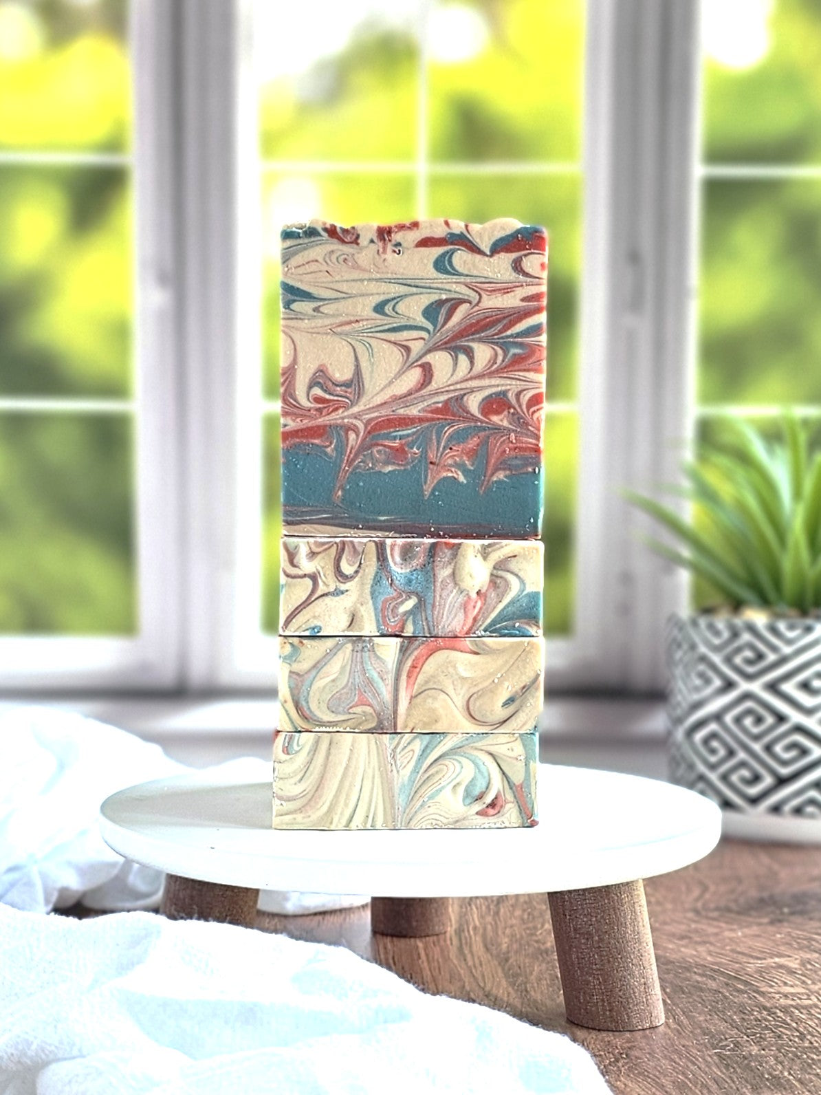 Four Amillia Acres ~ Goat Milk Soaps, featuring swirling patterns, are stacked vertically on a round wooden stand beside a potted plant against a white curtain, creating a natural and serene atmosphere. The Barbershop scents of these nourishing soaps enhance the peaceful setting.