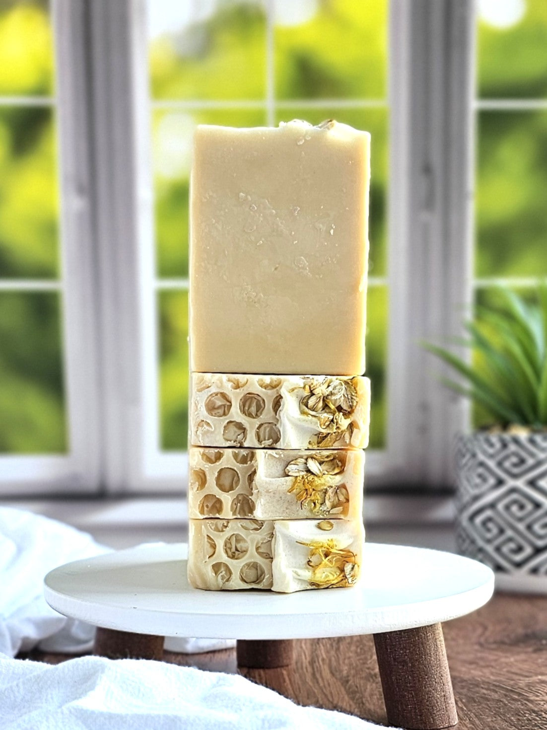 A stack of Amillia Acres Nourish Goat Milk Soaps is displayed on a small wooden stand. The handmade artisanal soaps, crafted with natural ingredients, boast a honeycomb texture and are garnished with dried flowers. A blurred window and plant in the background enhance the natural ambiance.