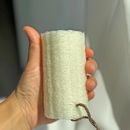 A hand holds the Amillia Acres Natural Loofah Sponge, featuring a brown string, against a softly lit background.