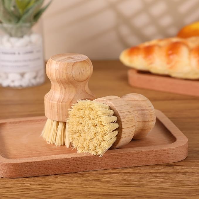 Two Amillia Acres Dish Scrubber brushes with wooden handles and durable bristles rest on a wooden tray, with blurred bakery items and a plant in the background.