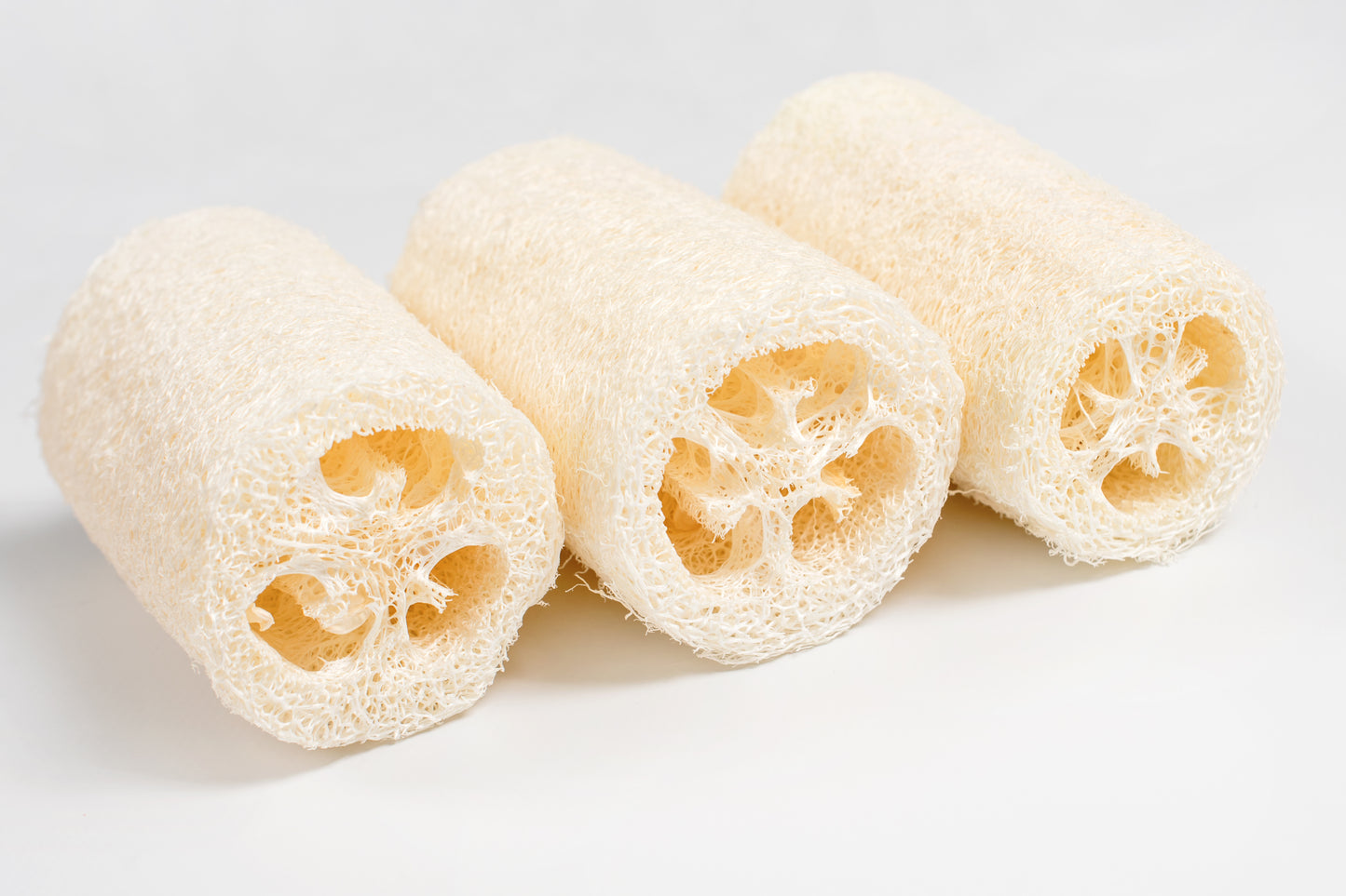 Three Amillia Acres Natural Loofah Sponges are arranged side by side on a white surface, highlighting their fibrous texture and hollow centers, ideal for gentle exfoliation as an eco-friendly scrubber.