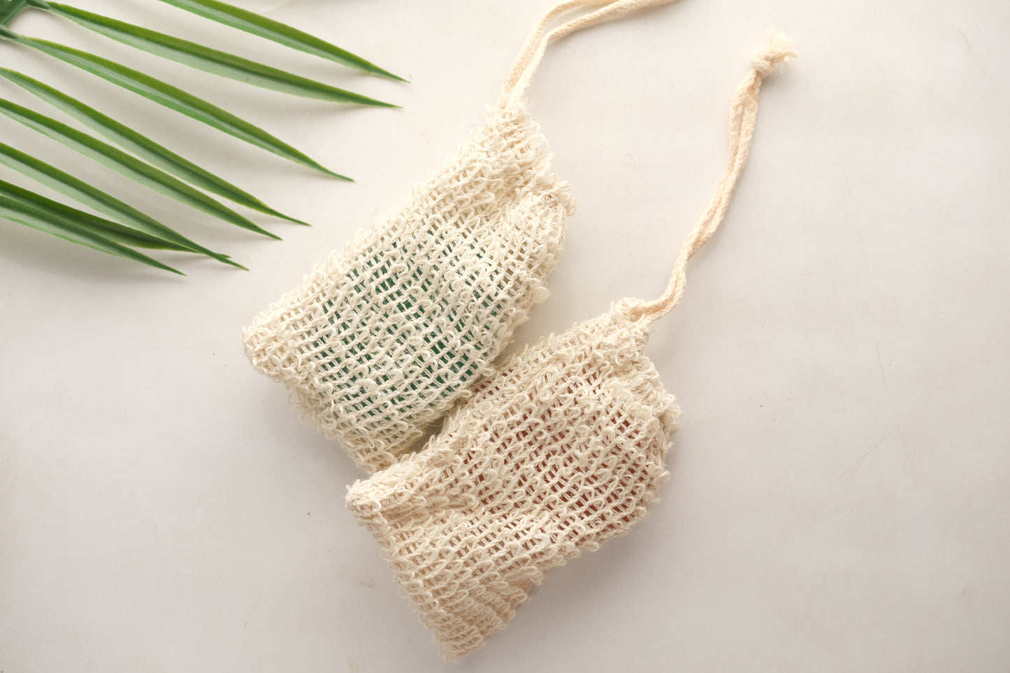 Two Amillia Acres Exfoliating Soap Saver Bags in natural woven mesh with drawstrings are displayed on a light surface, with a green palm leaf visible in the top left corner.