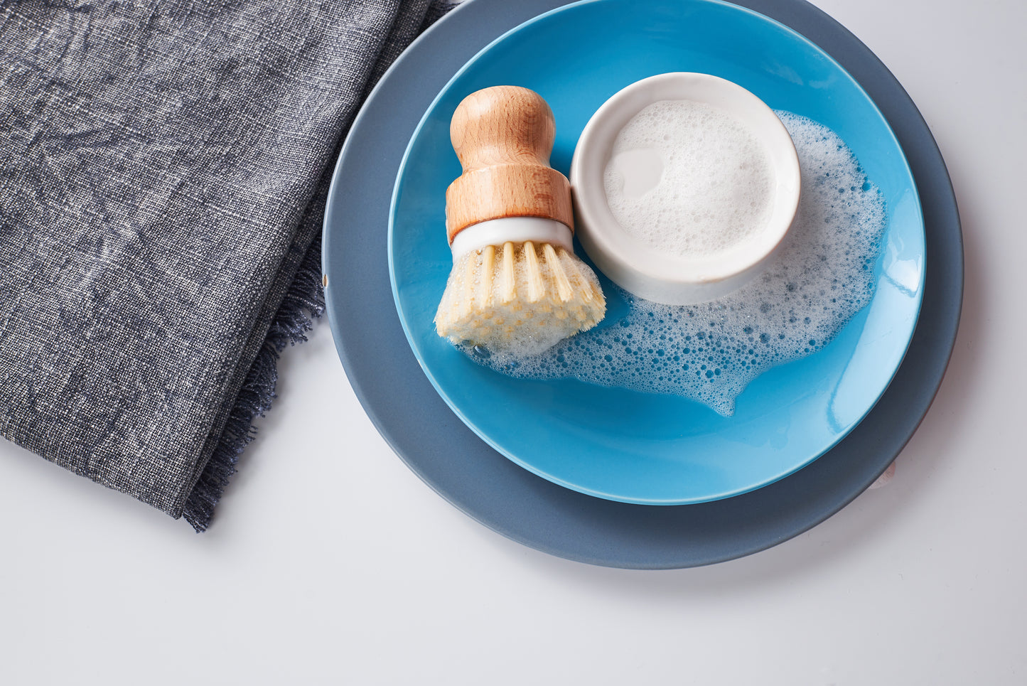 The Amillia Acres Dish Scrubber with durable bristles rests on a blue plate next to a bowl of soapy foam and bubbles, accompanied by a gray textured cloth on a white surface.