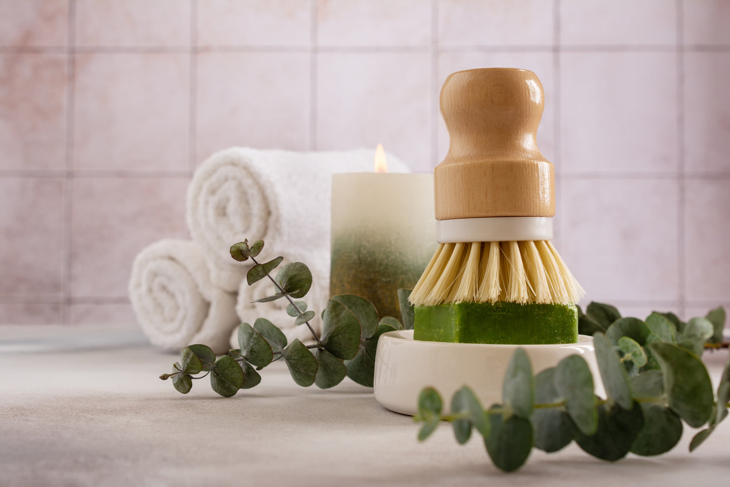 The Amillia Acres Dish Scrubber rests on a green soap bar in a dish, with eucalyptus leaves, rolled white towels, and a lit candle in the background on a light surface.