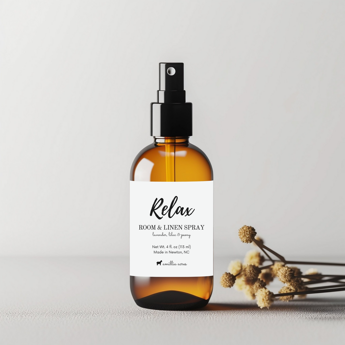 A brown glass spray bottle labeled "Room + Linen Spray" from Amillia Acres ~ Goat Milk Soaps and Lotions sits with dried flowers. The minimalist label highlights lavender and blue spruce notes, offering a soothing, elegant spa experience on a light surface.