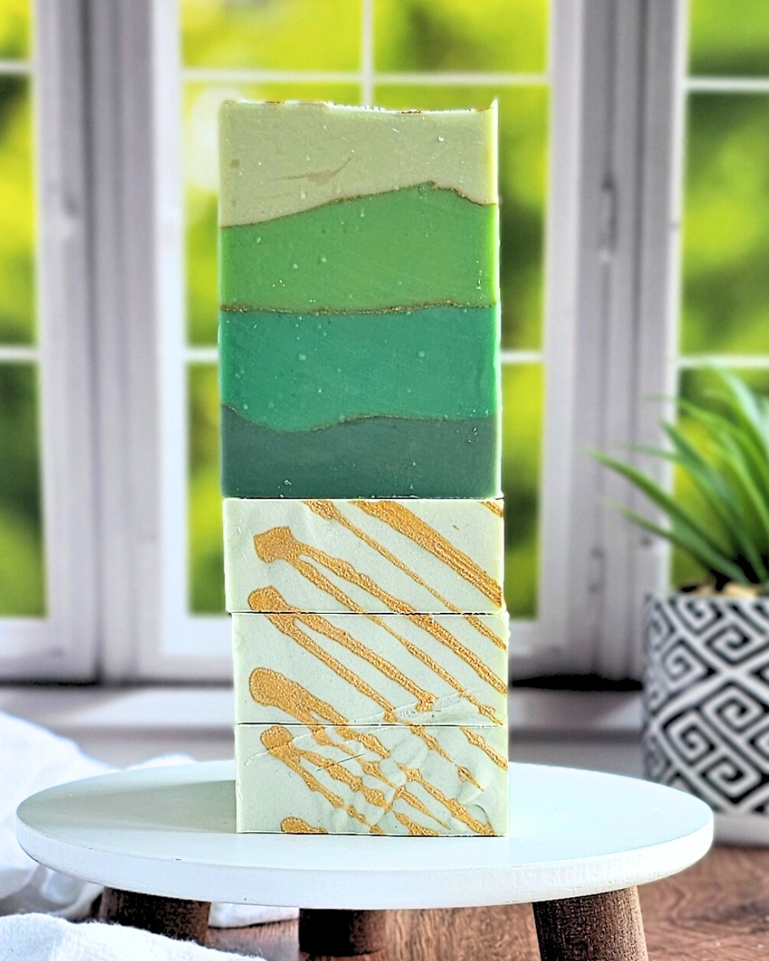 Three bars of Amillia Acres' Emerald Goat Milk Soap, with green and teal layers, are stacked atop a gold swirl bar. They evoke Irish Spring's freshness, perched on a white stand by a window with a blurred green scene and a plant behind them.