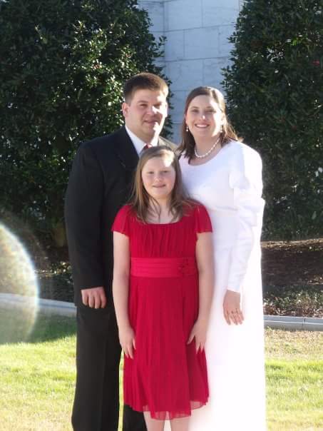 A man in a suit, a woman in a white dress, and a young girl in a red dress stand together smiling outside. They are surrounded by greenery, and sunlight creates a lens flare on the left.