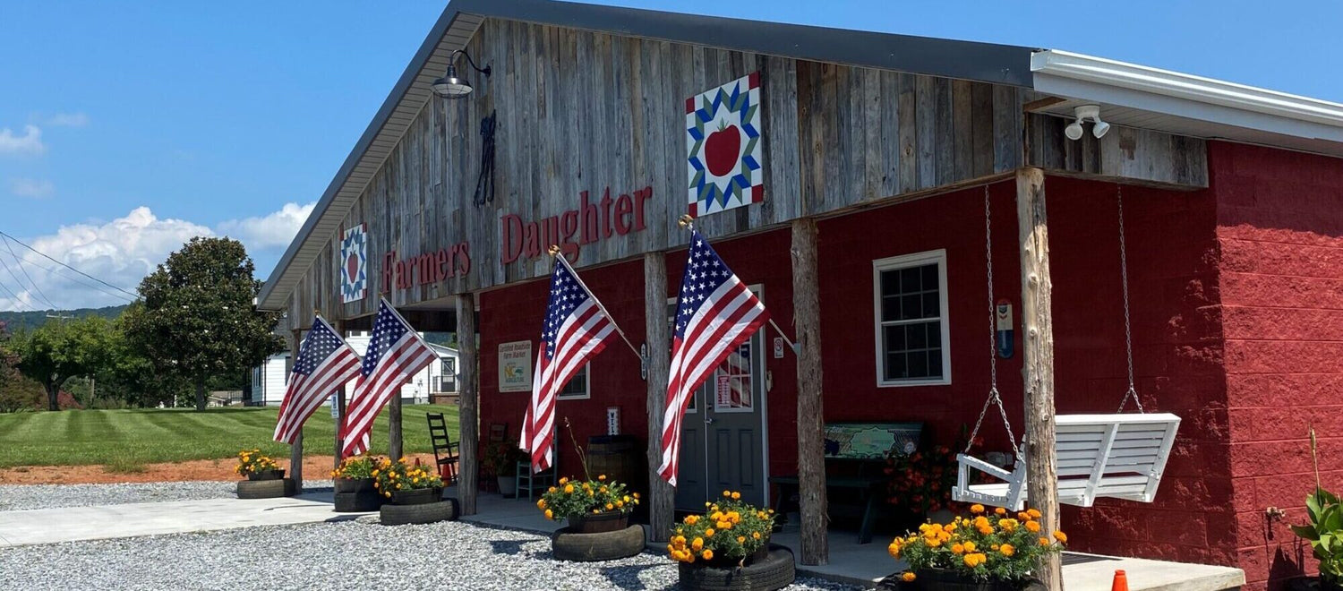 A rustic building with a wooden facade labeled "Farmer's Daughter." Multiple U.S. flags are displayed in front, alongside planters with vibrant orange flowers. A white swing hangs from the porch. The sky is clear and blue.