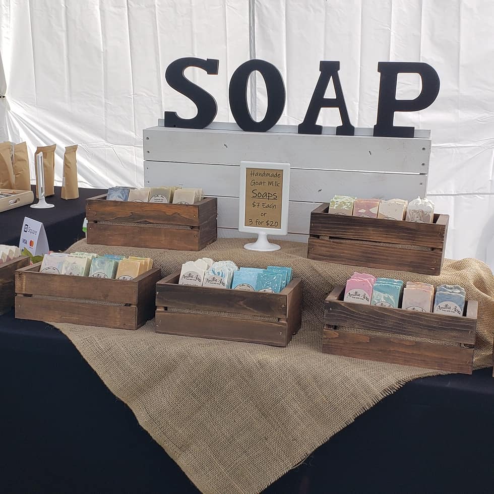 Display of handmade soaps in wooden crates on a table with a burlap cloth. A sign reads "Handmade Goat Milk Soaps $7 Each, 3 for $20." Large letters spelling "SOAP" are displayed on a white backdrop.